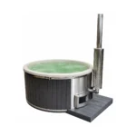 Ronde hottub met lichte kuip en water, donkergrijze verticale buitenbekleding, geïntegreerde roestvrijstalen kachel met glazen deur, hoge schoorsteen met geperforeerd hitteschild en donker opstapplatform op witte achtergrond.