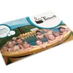 Schuin liggende TimberIN-productcatalogus met op de omslag een groep kinderen en volwassenen in een ronde houten hottub op witte achtergrond.