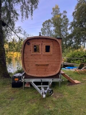 Vooraanzicht van de TimberIN Nordic Square mobiele sauna op aanhanger aan een meer