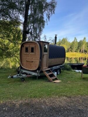 Zijaanzicht van de TimberIN Nordic Square mobiele sauna op aanhanger met schoorsteen aan een meer