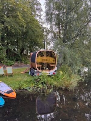 Achteraanzicht met panoramisch glas van de TimberIN Nordic Square mobiele sauna op aanhanger aan een meer