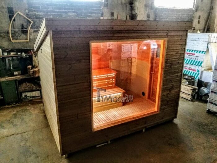 Compacte vierkante houten buitensauna met groot voorraam en verlichte banken in een werkplaats