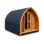 Boogvormig houten glampingpod met donker shingledak, open houten deur met smal raam, overdekte veranda en houten vloer op witte achtergrond.