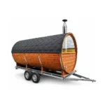 Mobiele tonvormige houten buitensauna op een dubbelassige aanhangwagen met donker shingledak, gebogen panoramisch voorraam, roestvrijstalen schoorsteen en dissel op witte achtergrond.