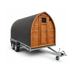 TimberIN Nordic Mobile Sauna Pod gebogen houten podsauna met zwarte shinglebekleding, houten voorgevel en deur met raam, gemonteerd op een tweeassige aanhanger met dissel op witte achtergrond.