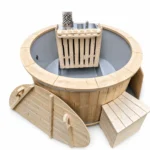 Compacte ronde houten hottub met grijze kuip, interne roestvrijstalen houtkachel achter een houten beschermrek, schoorsteen met geperforeerd hitteschild, losse tweedelige houten afdekking en houten opstap op witte achtergrond.