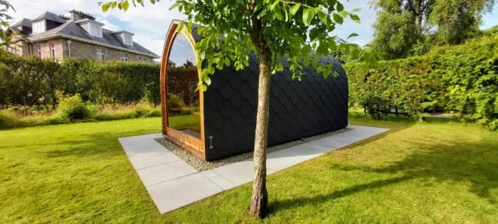 Donkere compacte podsauna met glazen voorkant tussen tuinplanten