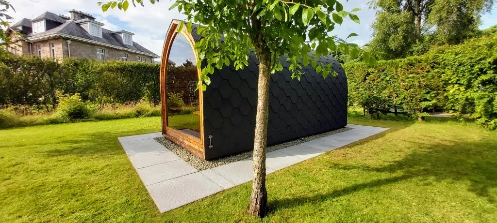 Donkere compacte podsauna met glazen voorkant tussen tuinplanten