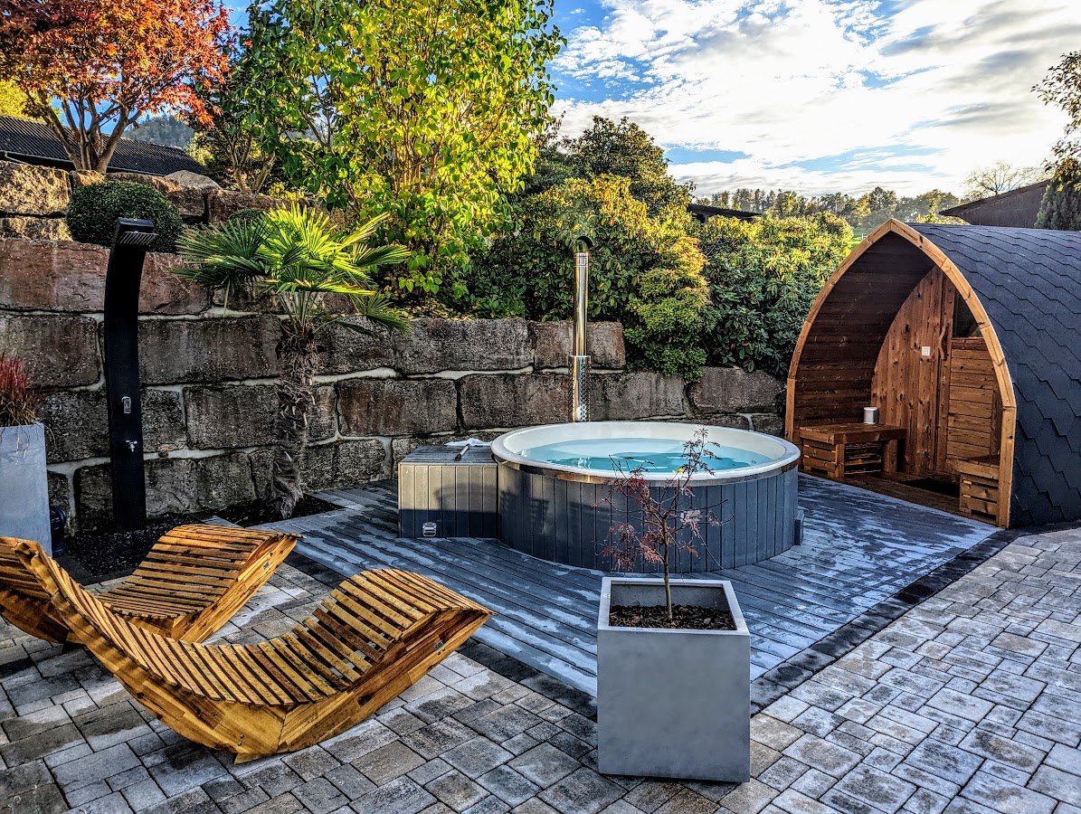 Ronde hottub op een stenen tuinterras met houten ligstoelen, groen en een podsauna ernaast